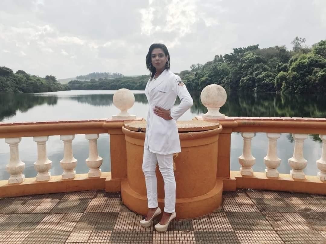 Foto de uma profissional de saúde vestindo um jaleco branco e calça branco. Ela está com uma das mãos na cintura. A enfermeira está em frente a um balaústre laranja e branco. Atrás, está uma paisagem, com um lago e árvores em volta.